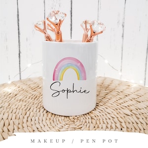 May include: White ceramic pen pot featuring a watercolour rainbow and the name "Sophie". It contains rose gold pens with diamond-shaped tops. The words "MAKEUP / PEN POT" are at the bottom. The pot sits on a woven mat.