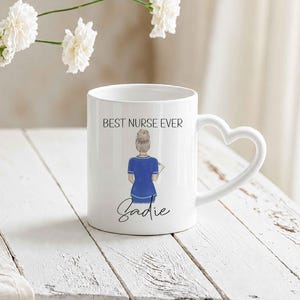 May include: White ceramic mug with a heart-shaped handle. The mug features the text "BEST NURSE EVER" and an illustration of a nurse in blue scrubs. The name "Sadie" is written below the illustration. The mug is on a white wooden surface.