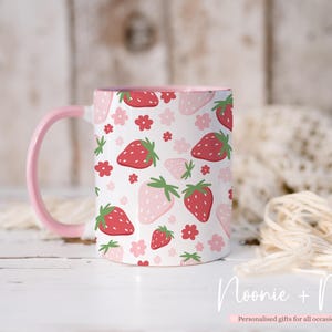 May include: A white ceramic mug with a pink handle and interior. The mug is decorated with a pattern of red and pink strawberries and small pink flowers. The text "Noonie + Me" is visible.