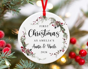 Può includere: Una decorazione rotonda in ceramica bianca con un nastro rosso. Il design presenta una ghirlanda ad acquerello con fogliame verde e bacche rosse che circonda il testo nero: FIRST Christmas AS AMELIA'S Auntie & Uncle 2026 in vari caratteri.