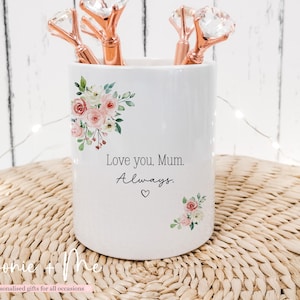 May include: White ceramic pen holder with floral accents and the text "Love you, Mum. Always." Includes rose gold diamond-topped pens. The pen holder is decorated with pink and white flowers and green leaves.