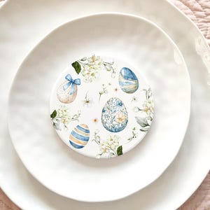 May include: A white ceramic coaster with a blue and white floral design featuring eggs with blue and white patterns. The coaster is on a white plate.