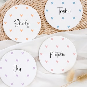 May include: Four round white coasters with different coloured hearts and names written in black. The names are Shelly, Trisha, Natalie, and Joy.
