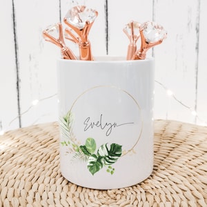 May include: White ceramic pencil holder with a gold circle and the name "Evelyn" written in cursive. The holder has a tropical leaf design and is filled with rose gold pens with diamond-shaped tops.