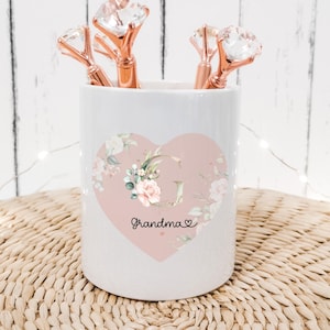 May include: White ceramic pencil holder with a pink heart design. The heart has a floral pattern and the word "Grandma" written on it. The holder has rose gold diamond-shaped pens inside.