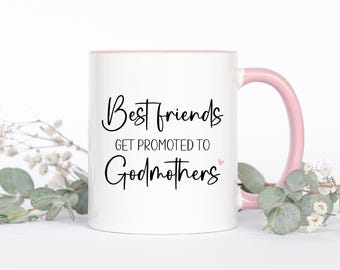 May include: A white ceramic mug with a light pink handle and interior rim. The front features black cursive and sans-serif text that reads Best friends GET PROMOTED TO Godmothers with a small pink heart icon next to the word Godmothers.
