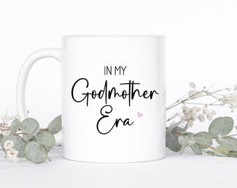 May include: A white ceramic mug featuring the black printed text "IN MY Godmother Era" with a small pink heart icon. The mug has a standard handle and a smooth, glossy finish, presenting a clean and simple design for a beverage container.