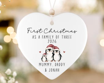May include: A white heart-shaped ceramic ornament hanging from a string with a wooden bead. It features the text First Christmas AS A FAMILY OF THREE 2026, an illustration of three penguins wearing Santa hats, and the text MUMMY, DADDY & JONAH.