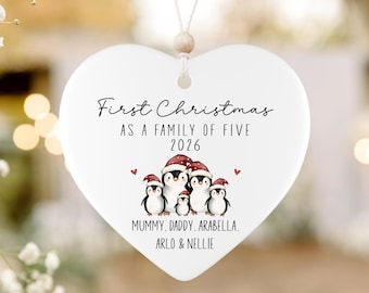 May include: A white heart-shaped ceramic ornament hanging from a string with a wooden bead. It features a graphic of five penguins wearing red Santa hats, with the text "First Christmas AS A FAMILY OF FIVE 2026" and "MUMMY, DADDY, ARABELLA, ARLO & NELLIE".