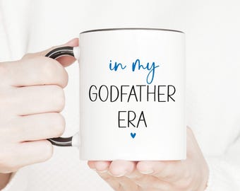 May include: White ceramic mug with a black rim and handle. The mug has blue text that reads "in my GODFATHER ERA" with a small blue heart below the text.