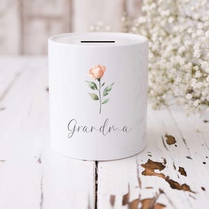 May include: White ceramic money bank with a slot on top. The bank is decorated with a watercolor-style peach rose and the word "Grandma" in script. The bank is cylindrical and sits on a white wooden surface.
