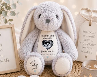 May include: A plush gray rabbit toy with long ears and a cream-colored belly patch. The patch features a black and white ultrasound image, the text "HELD IN OUR hearts forever", and the name "Daniel James Holder". The left foot displays "24th July 2026".