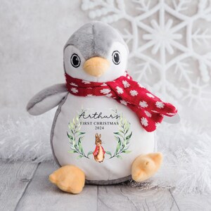 May include: A gray and white penguin plush toy wearing a red and white polka dot scarf. The penguin has a white belly with the text "Arthur's First Christmas 2024" and a green wreath with a rabbit in a red jacket.