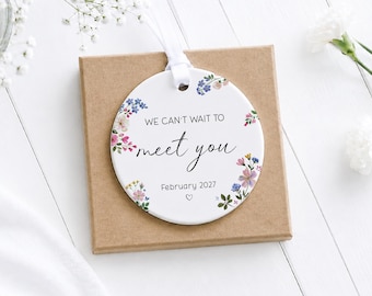May include: A round white ceramic ornament featuring a floral border with blue, pink, and green flowers. The text reads WE CAN'T WAIT TO meet you, followed by February 2027 and a small heart symbol. A white ribbon is attached to the top of the ornament.