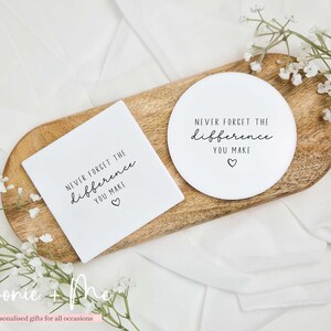 May include: A wooden serving tray holds a white square coaster and a round coaster. Both coasters have the text "NEVER FORGET THE difference YOU MAKE" with a heart symbol. The tray is surrounded by white fabric and sprigs of white flowers.