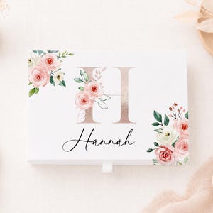 May include: A rectangular white box featuring a large rose gold letter H and the name Hannah in black script. The design is decorated with watercolour-style pink and white roses with green leaves in the top left and bottom right corners.