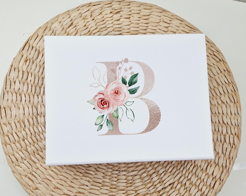 Puede incluir: Una impresi&oacute;n en lienzo blanco con una letra "B" de oro rosa decorada con flores de acuarela rosa y verde.