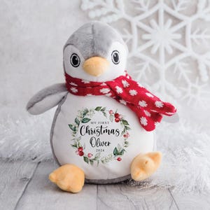 May include: A gray and white penguin plush toy wearing a red and white polka dot scarf. The penguin has a wreath with green leaves and red berries around its belly with the text "My First Christmas Oliver 2024".