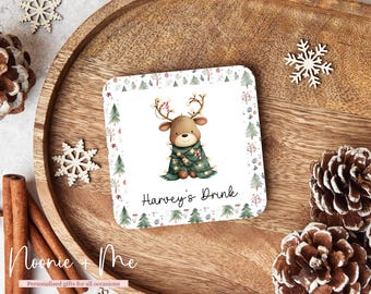 May include: Square Christmas coaster featuring a cartoon reindeer wearing a green plaid blanket and string lights. The coaster has a white background with a border of Christmas trees and the text "Harvey's Drink".