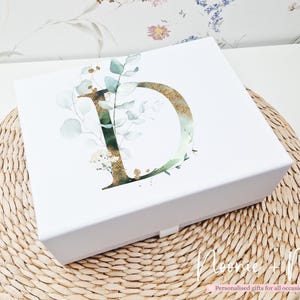 May include: A white gift box featuring a gold-glittered letter "D" and a watercolour eucalyptus and floral design. The box sits on a woven mat. The text "Noonie + Me" and "Personalised gifts for all occasions" is visible.