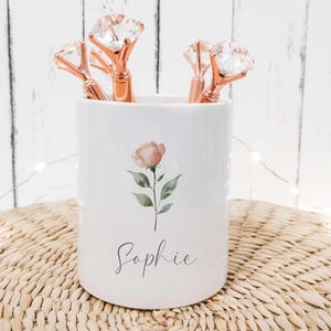 May include: A white ceramic pen pot decorated with a watercolour rose and the name "Sophie". It holds rose gold pens with diamond-shaped tops. The pot sits on a woven mat, with fairy lights in the background.