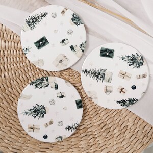 May include: Three white ceramic coasters with a winter holiday theme. Each coaster features a different design with green trees, white gift boxes, and white snowflakes on a white background.