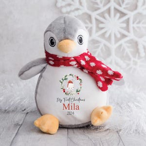 May include: A gray and white penguin plush toy wearing a red and white polka dot scarf. The penguin has a wreath with a Santa hat on its chest and the text "My First Christmas Mila 2024".