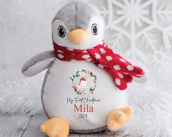 May include: A gray and white penguin plush toy wearing a red and white polka dot scarf. The penguin has a wreath with a Santa hat on its chest and the text "My First Christmas Mila 2024".
