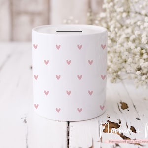 May include: White ceramic money box adorned with small pink hearts. The cylindrical money box has a slot at the top. It is a decorative item, sitting on a white wooden surface. The money box is a decorative item.