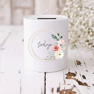 May include: White ceramic money box with a gold circle design and watercolour floral accents. The name "Evelyn" is written in black script inside the circle.