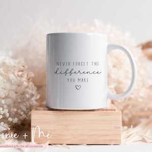 May include: White ceramic mug with the text "NEVER FORGET THE difference YOU MAKE" in black script and a small heart. The mug is on a small wooden block, with a soft, blurred background of white and beige flowers.