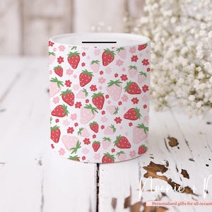 May include: A white ceramic money box with a slot on top, decorated with a pattern of red and pink strawberries and small red flowers on a white background. The bank is cylindrical and sits on a white wooden surface.