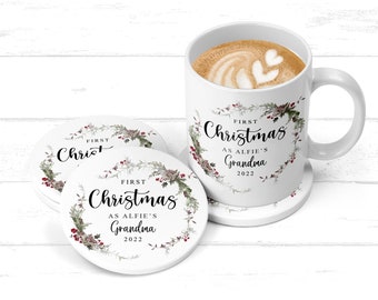 May include: A white ceramic mug and coasters set. The mug and coasters feature the text "First Christmas as Alfie's Grandma 2022" surrounded by a wreath of green leaves, red berries, and gray succulents. The mug contains a coffee drink with heart-shaped foam art.