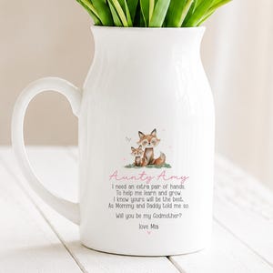 May include: White ceramic jug with a handle, decorated with a watercolour illustration of two foxes and the text "Aunty Amy". The jug has a curved handle and contains green stems.