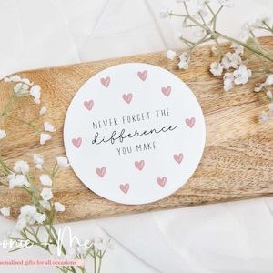 May include: A white, circular coaster with pink hearts and the text "NEVER FORGET THE difference YOU MAKE." The coaster is on a wooden tray, decorated with white flowers.