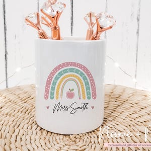 May include: White ceramic pencil pot featuring a pastel rainbow design, an apple, and the name "Miss Smith". It holds three rose gold pens with diamond-shaped tops. The pot sits on a woven surface.
