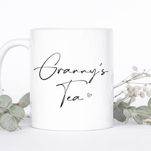 May include: A white ceramic mug featuring the black cursive text Granny's Tea with a small heart symbol. The mug has a standard handle and a smooth finish, presenting a clean design suitable for holding hot beverages in a home or office setting.