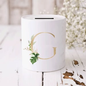 May include: White ceramic money box with a gold letter G and green and white floral design.