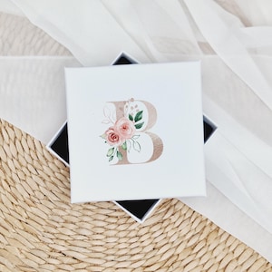 May include: A white gift box featuring a gold letter "B" and pink watercolour flowers. The box is resting on a woven straw surface.
