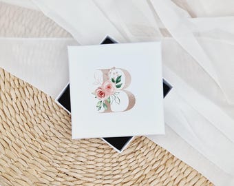 May include: A white gift box featuring a gold letter "B" and pink watercolour flowers. The box is resting on a woven straw surface.