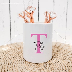 May include: White ceramic pencil holder with a pink letter T and the name Tilly written in black cursive. The holder is filled with rose gold pens with clear crystal tops.