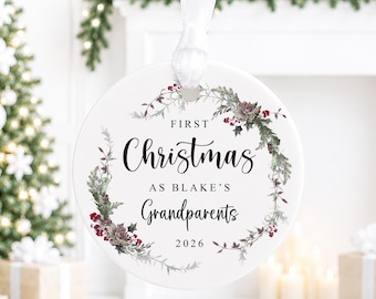 May include: A round white ceramic Christmas ornament featuring a wreath of green foliage and red berries. The text reads: FIRST Christmas AS BLAKE'S Grandparents 2026. A white satin ribbon is attached to the top for hanging the circular decoration.