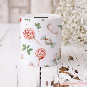 May include: White ceramic money box with a slot on top, decorated with watercolour illustrations of red and white swirl lollipops, wrapped sweets, and green leaves. The money box is cylindrical and sits on a white wooden surface.