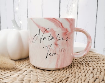 May include: A white and pink marble-patterned ceramic mug with the words "Natalie's Tea" in black script. A small heart is next to the word "Tea". The mug has a matching pink handle.
