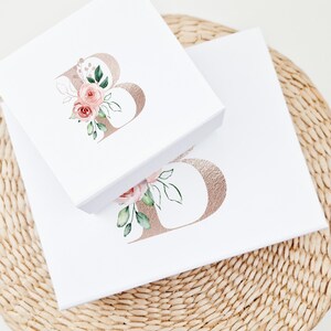 Puede incluir: Dos cajas de regalo blancas con tapas, cada una con una letra "B" de l&aacute;mina dorada con acentos florales de acuarela rosa. Las cajas est&aacute;n apiladas una encima de la otra.