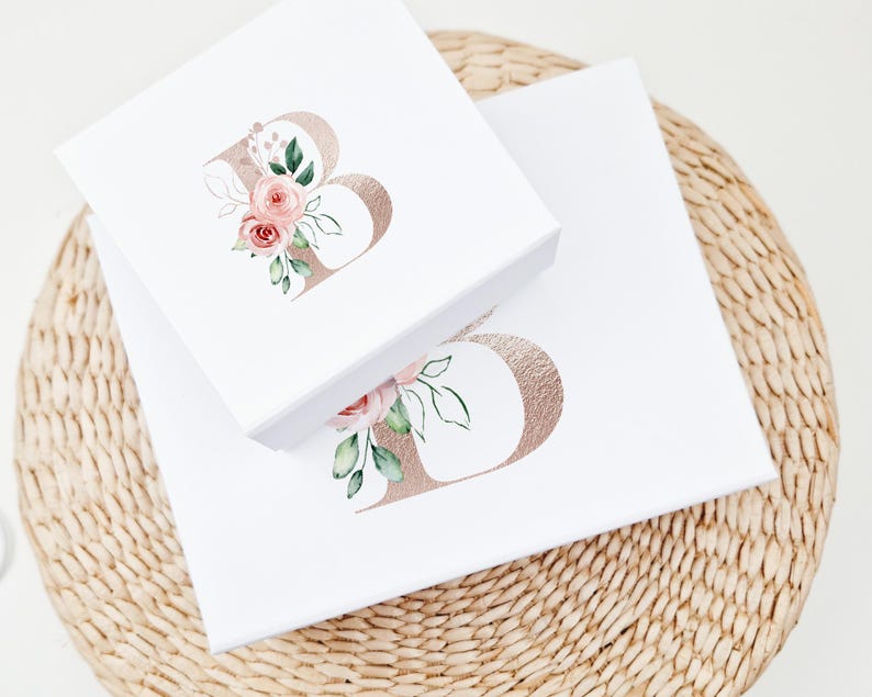 Puede incluir: Dos cajas de regalo blancas con tapas, cada una con una letra "B" de l&aacute;mina dorada con acentos florales de acuarela rosa. Las cajas est&aacute;n apiladas una encima de la otra.