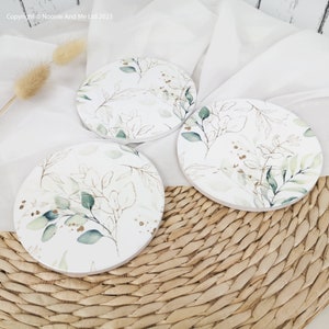 May include: Three round white coasters featuring a watercolour design of green leaves and gold accents. The coasters are displayed on a woven wicker surface.