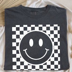 May include: Dark gray t-shirt featuring a white smiley face design set against a checkered background. The smiley face has a simple, curved mouth and two oval eyes. The shirt is made of a soft material.