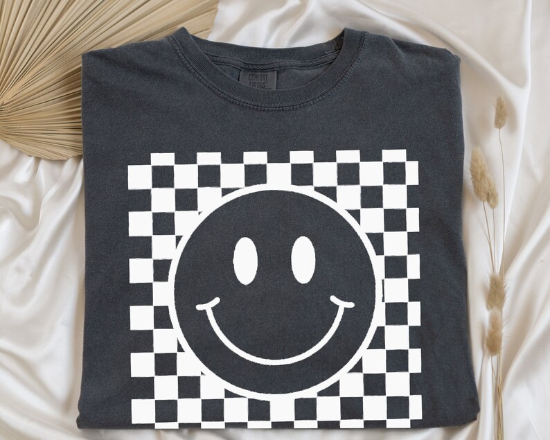 May include: Dark gray t-shirt featuring a white smiley face design set against a checkered background. The smiley face has a simple, curved mouth and two oval eyes. The shirt is made of a soft material.