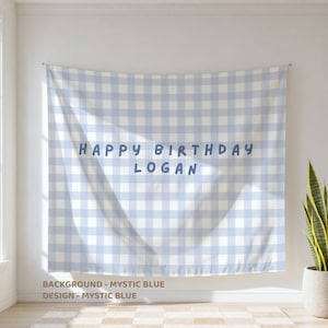 May include: A fabric wall tapestry featuring a light blue and white gingham check pattern. The text "HAPPY BIRTHDAY LOGAN" is printed in a dark blue, rounded sans-serif font across the center of the fabric, which hangs flat against a wall.
